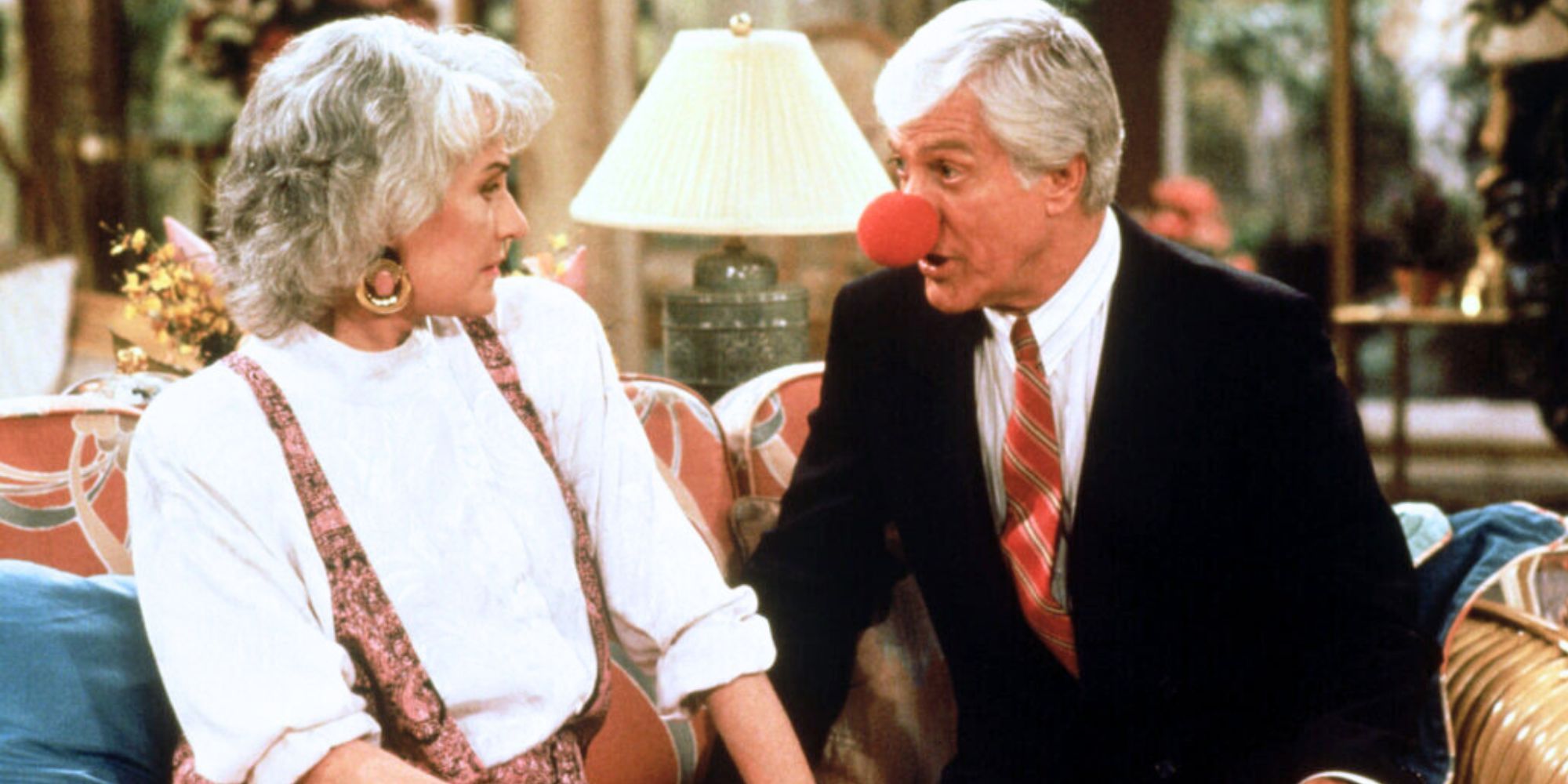 Bea Arthur looking at Dick Van Dyke sitting next to her on a couch while he has a clown nose on in The Golden Girls