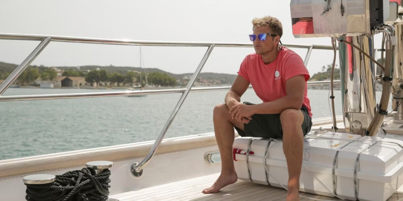 Tom Pearson sitting on a container on the deck of a ship with water and a coastline in the distance