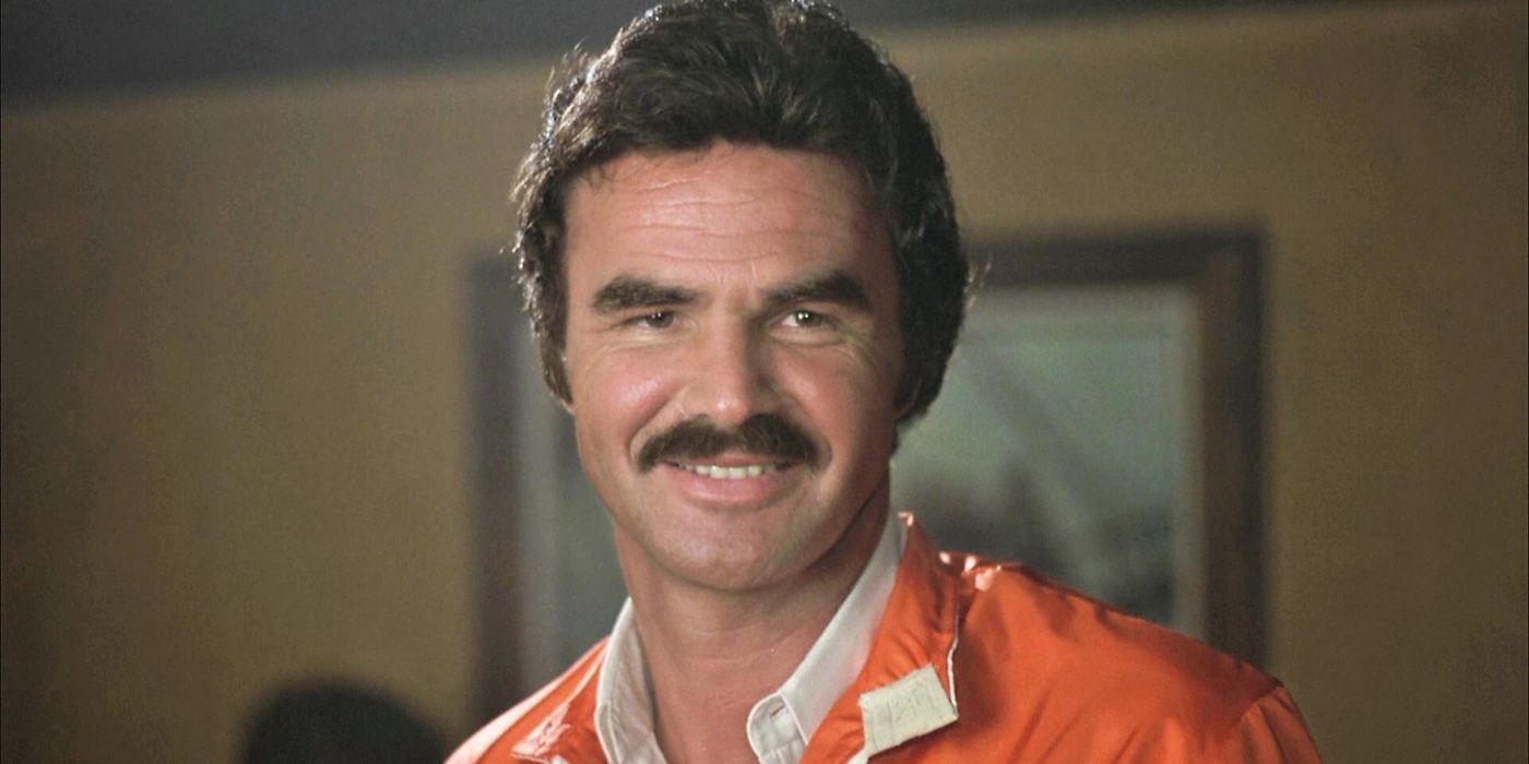 Burt Reynolds smiling toward the camera wearing an orang track jacket in The Cannonball Run