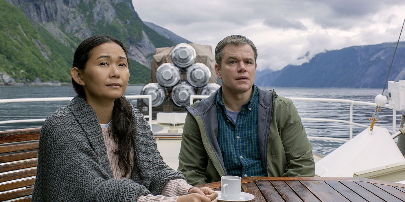 Hong Chau y Matt Damon se sientan en una mesa en un bote en un lago, mirando fuera de la cámara.