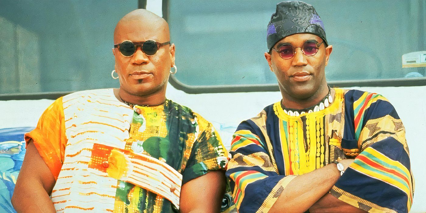 Two men wearing sunglasses lean against a car while wearing brightly colored African clothing