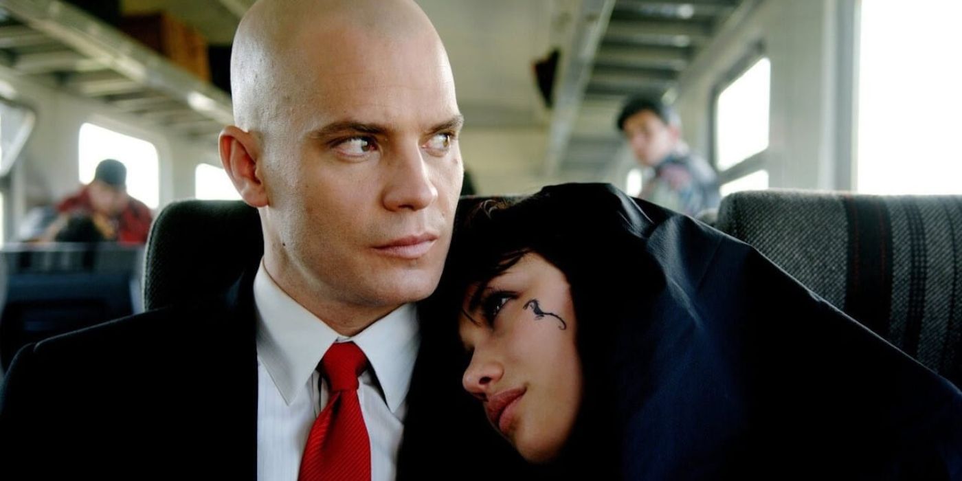 Agent 47 (Timothy Olyphant) sits on a train in his trademark suit and red tie while Nika Boronina (Olga Kurylenko) leans on him in 'Hitman' (2007).