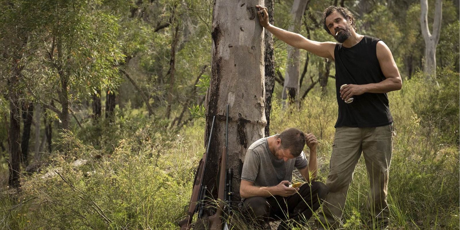 In the outback, a man sits against a tree, while another man stands nearby with a cigarette