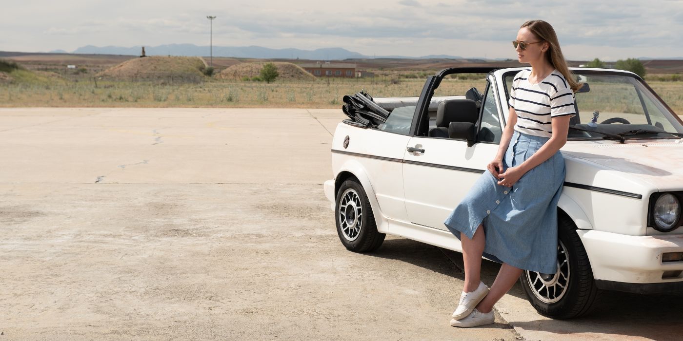 A woman in a jean skirt and stripped shirt leans against a white convertible in an empty parking lot