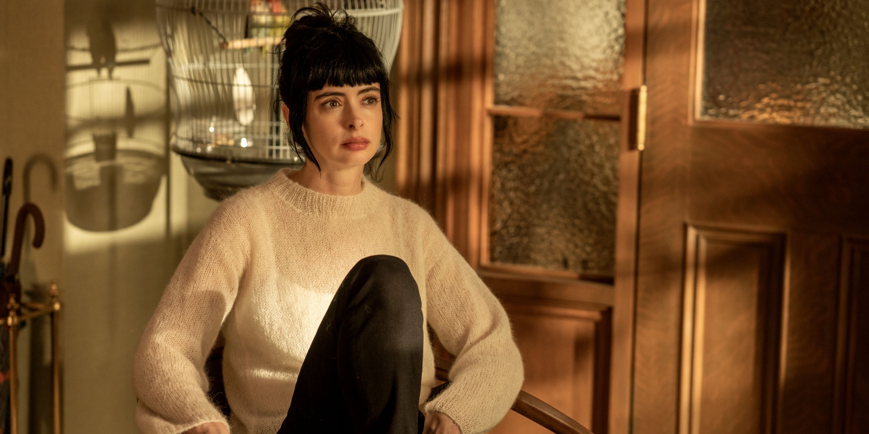 Krysten Ritter as Lucy sitting on a couch in a white sweater with her right knee bent in Orphan Black: Echoes