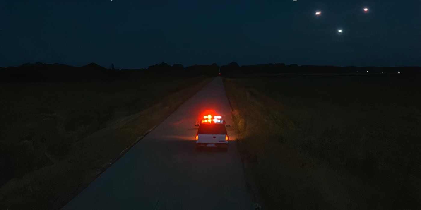 A police car drives down a country road as three lights appear in the distance