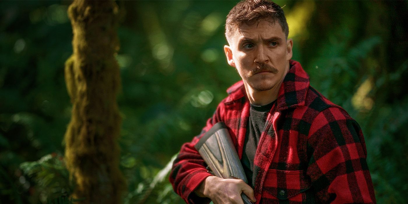 Kyle Gallner holding a rifle and frowning in the woods in a red plaid shirt