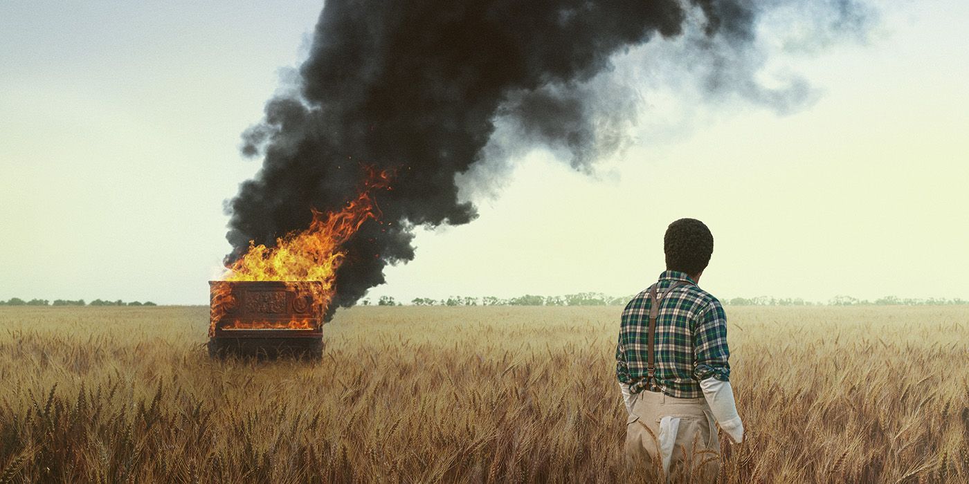 A man standing in a field with his back to the camera looking at a piano burning and billowing black smoke