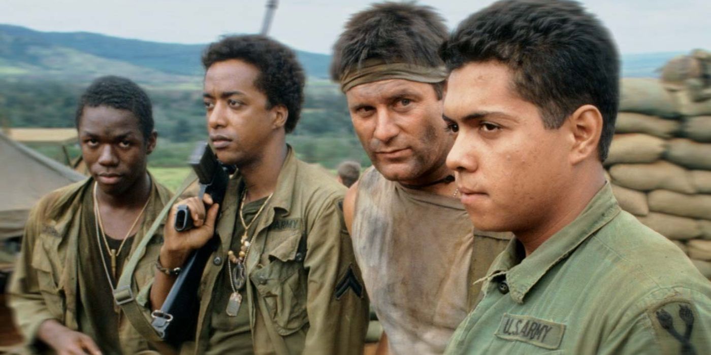 A group of soldiers stands in a line in Tour of Duty, one holds a gun over their shoulder and there are sandbags in the background.