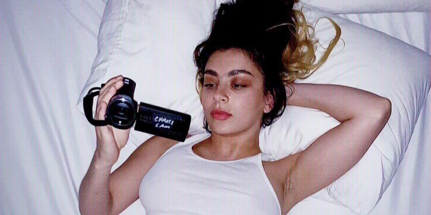 Charli XCX acostada en la cama y sosteniendo una videocámara en la mano