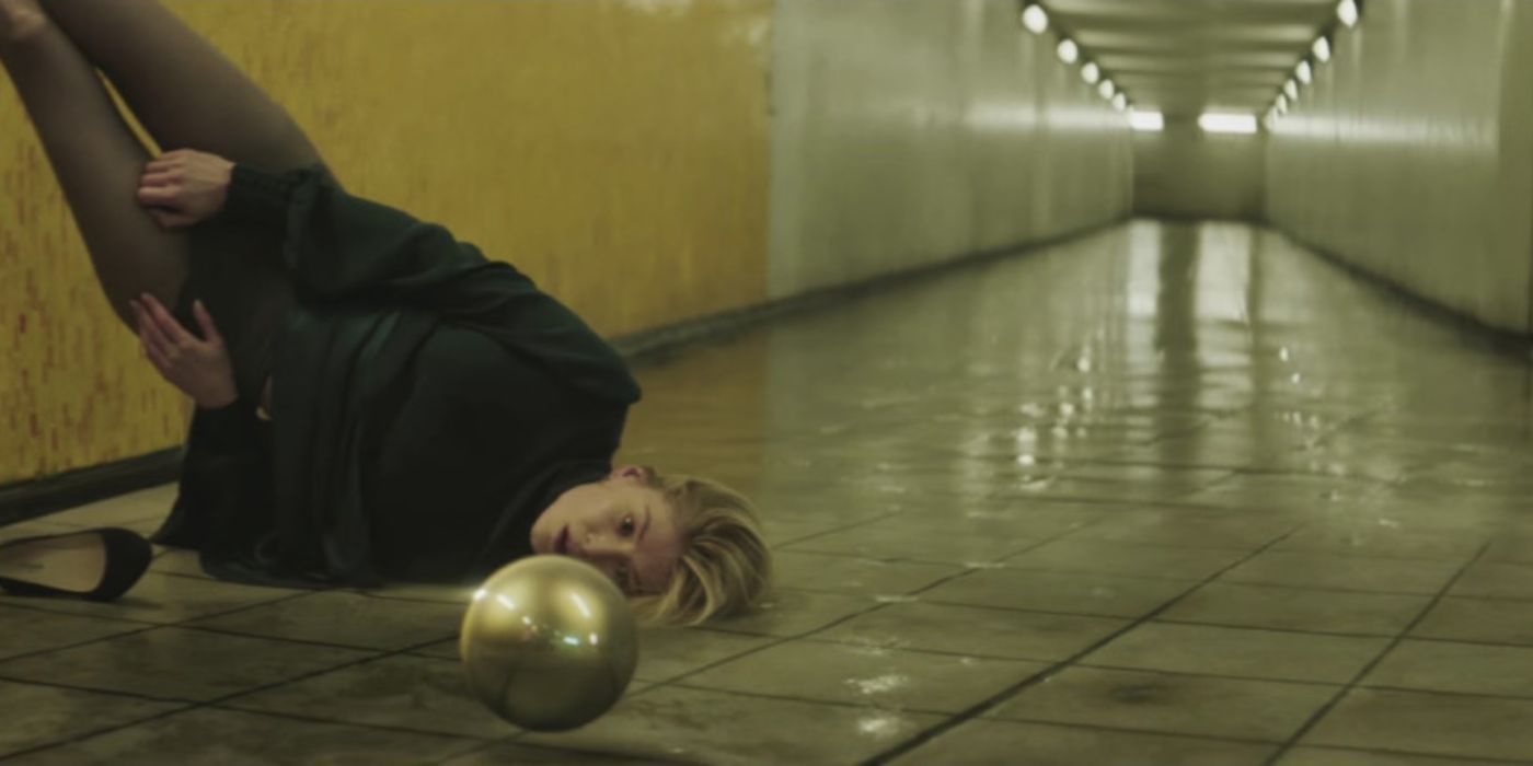 Rosamund Pike lays upside-down diagonally against a wall as a metal ball rolls by in the "Voodoo in my Blood' video