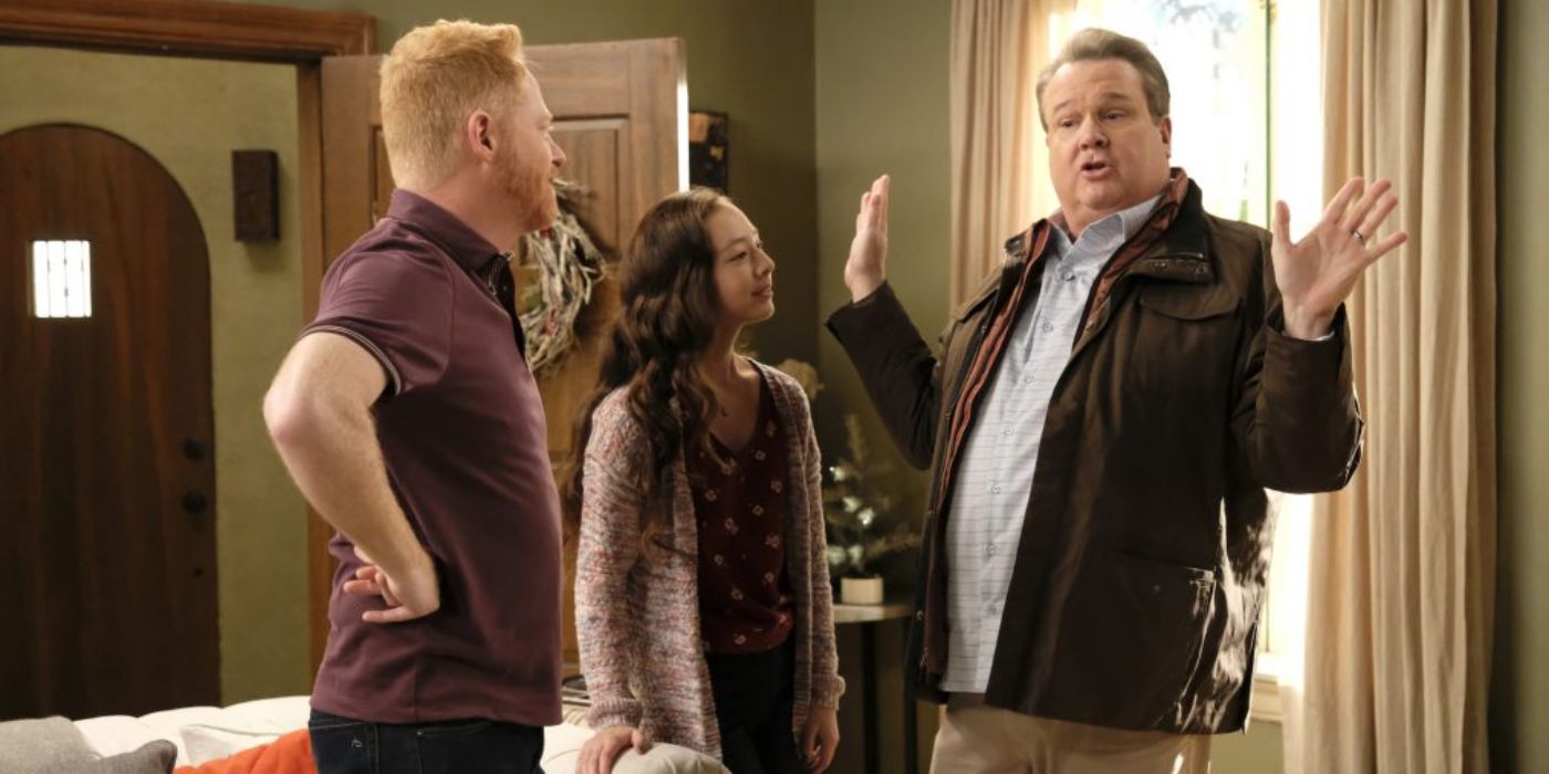 Cam gestures with his hands dramatically while Mitch stands with his hands on his hips and Lily talks with him in their living room on Modern Family.