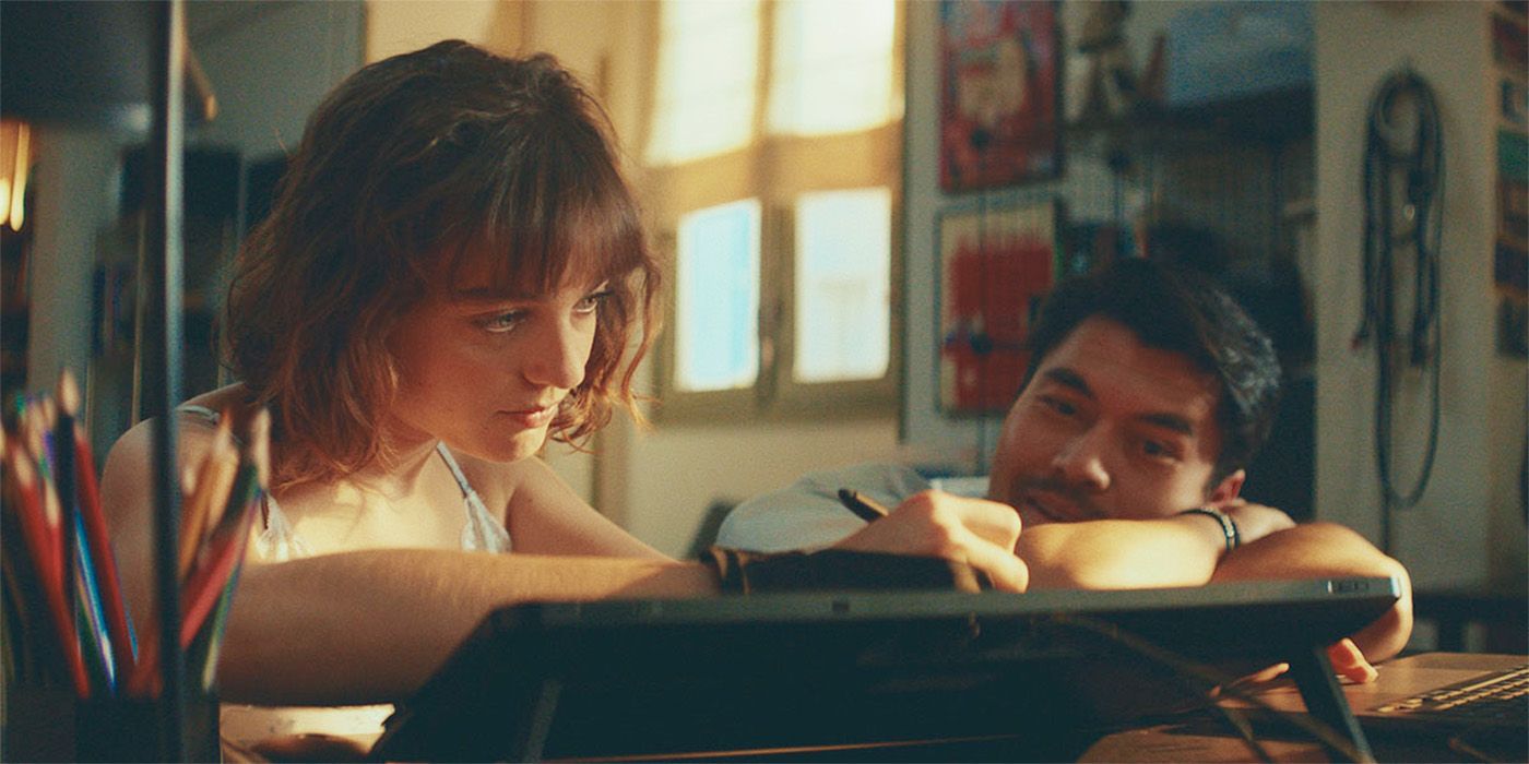 Henry Golding resting his head on a desk watching Beatrice Grannò sketch