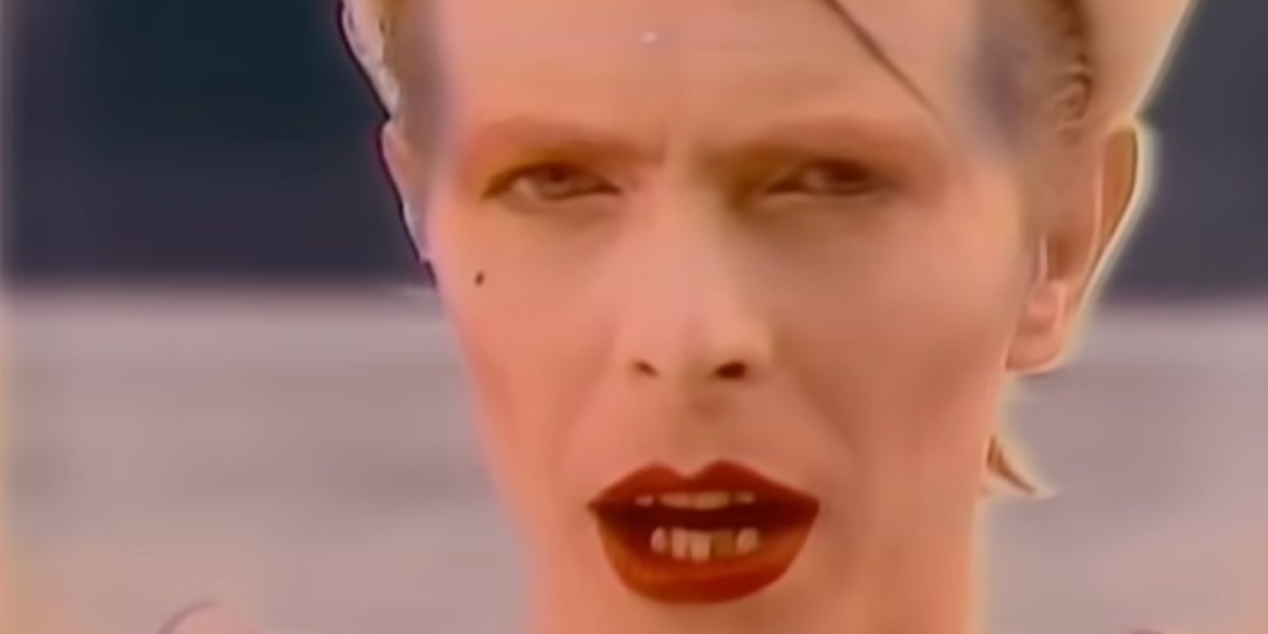 A closeup of David Bowie with red lipstick on and a line painted on his face in the music video Ashes to Ashes.