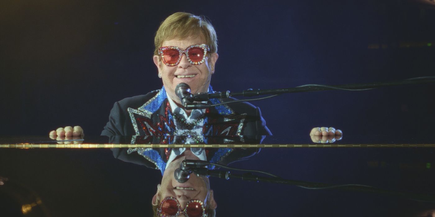 Elton John smiles through bejeweled glasses as he faces the grand piano on stage.