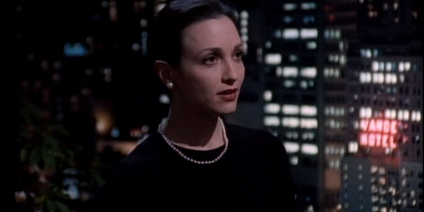 Lilith, wearing a black dress with pearls, stands in front of Frasier&rsquo;s living room window, which shows a vibrant city nightscape.