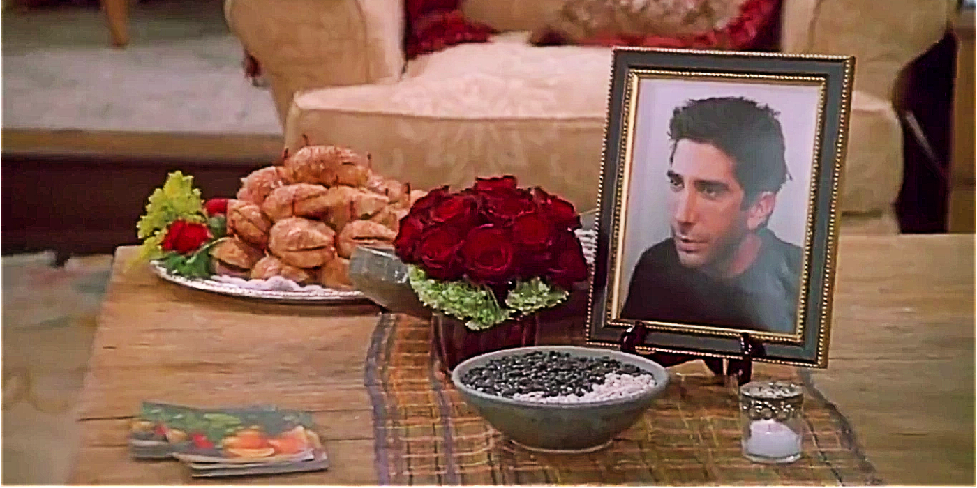 A screenshot from Ross Geller's fake memorial service in Friends, with a framed picture of Ross on the coffee table along with pastries and flowers