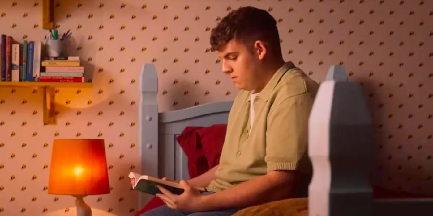 Isaac sits on a bed next to a lamp and holds a book in Heartstopper.