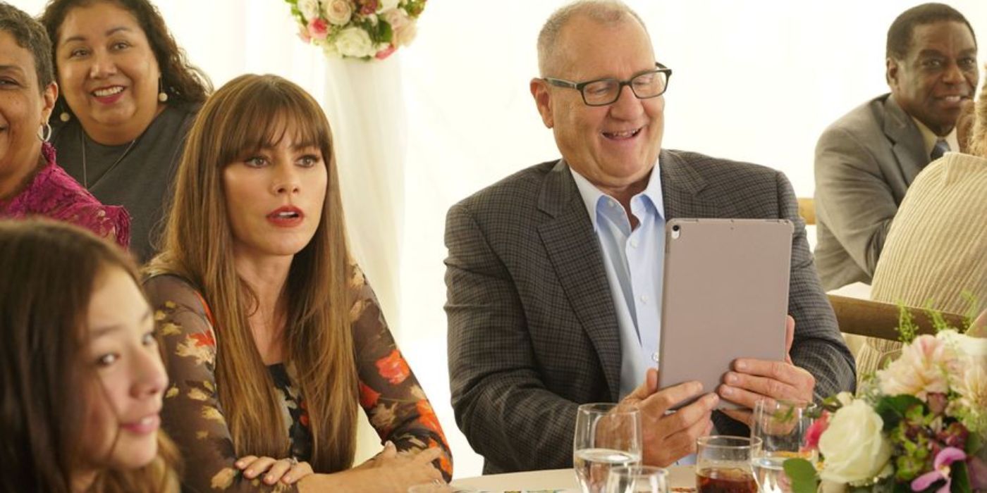 Jay looks amused at a tablet and Gloria looks surprised as they sit next to each other at a table surrounded by party guests at their 10th anniversary party in Modern Family.