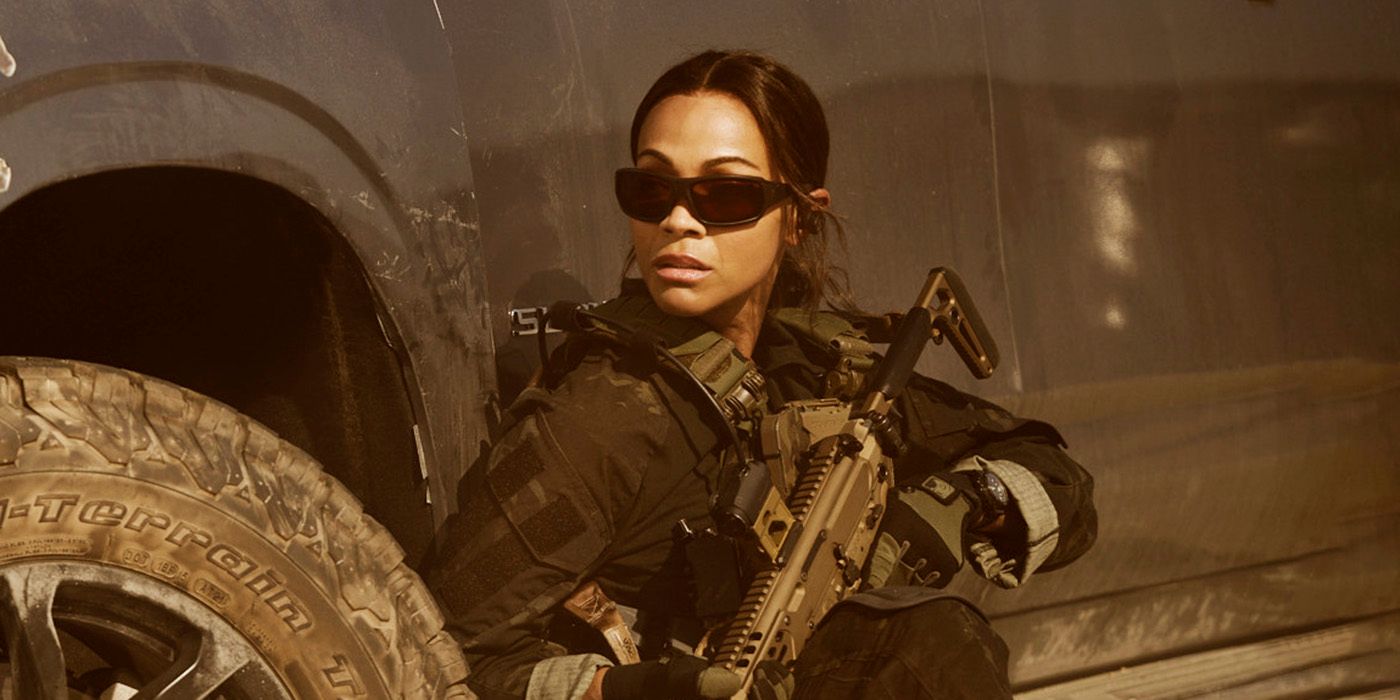 Zoe Saldana as Joe McNamara in military gear and sunglasses crouched behind the wheel well of a truck in 'Lioness' Season 2.