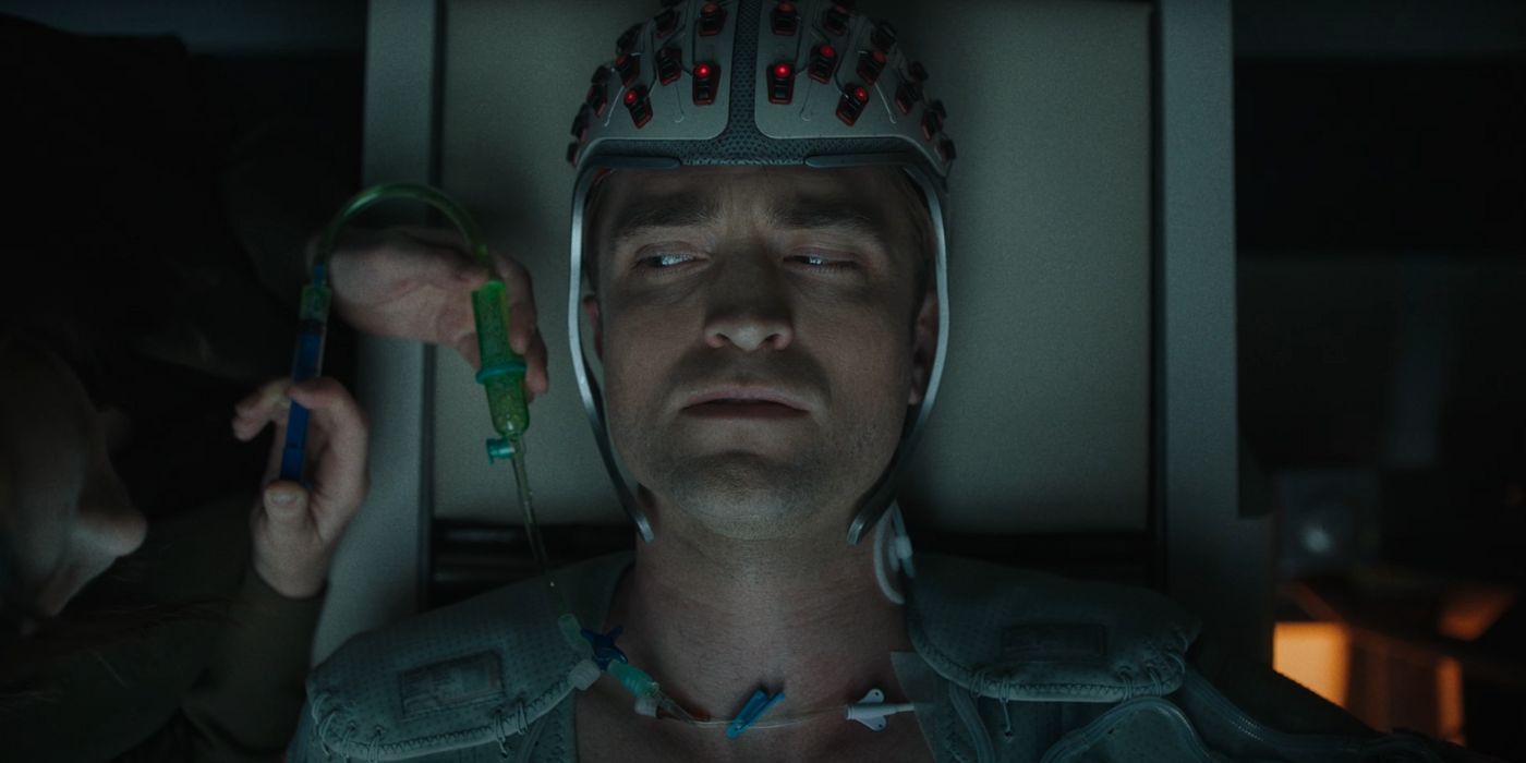 Robert Pattinson as Mickey looking confused wearing a helmet while fluid is pumped into him through IV.