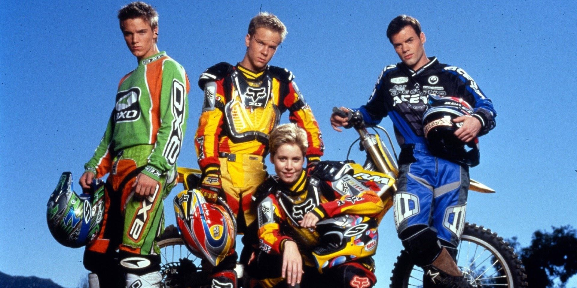 The cast of Motocrossed wearing biker gear and holding their biker helmets while leaning on their dirt bikes.