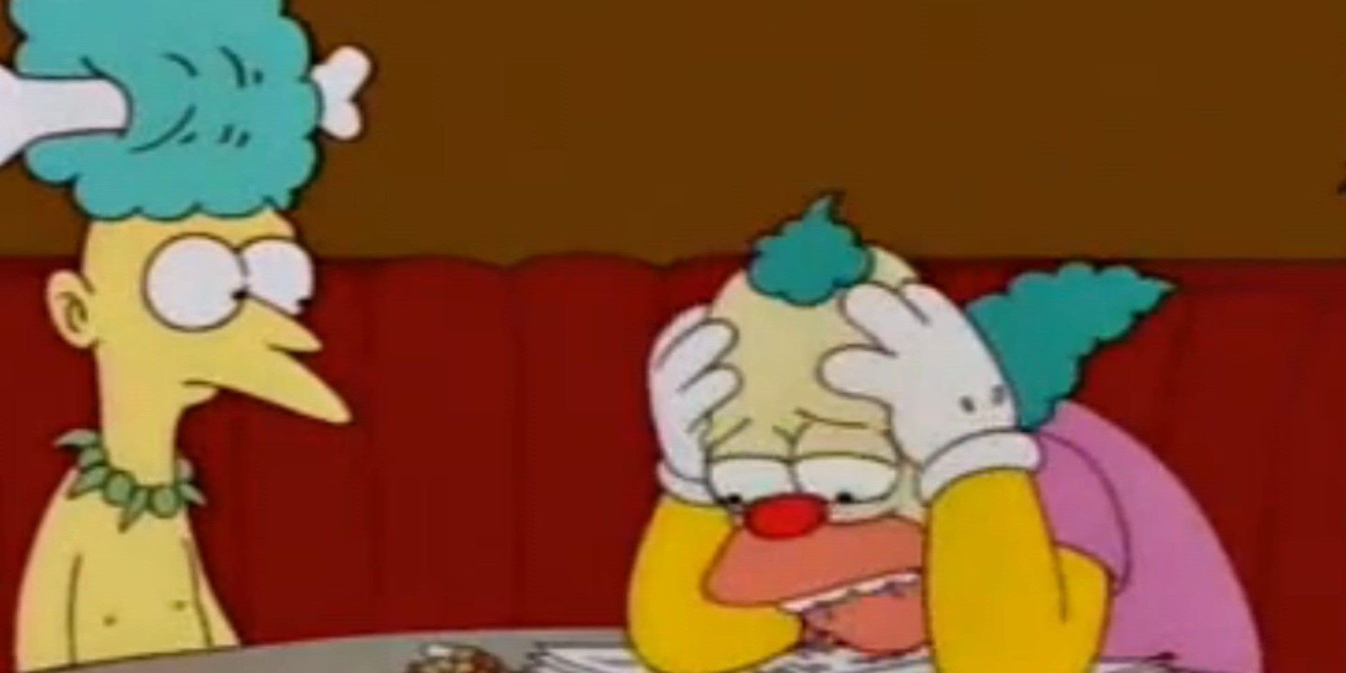 Krusty looks sad, holding his head in his hands as he shares a booth with another character on The Simpsons.