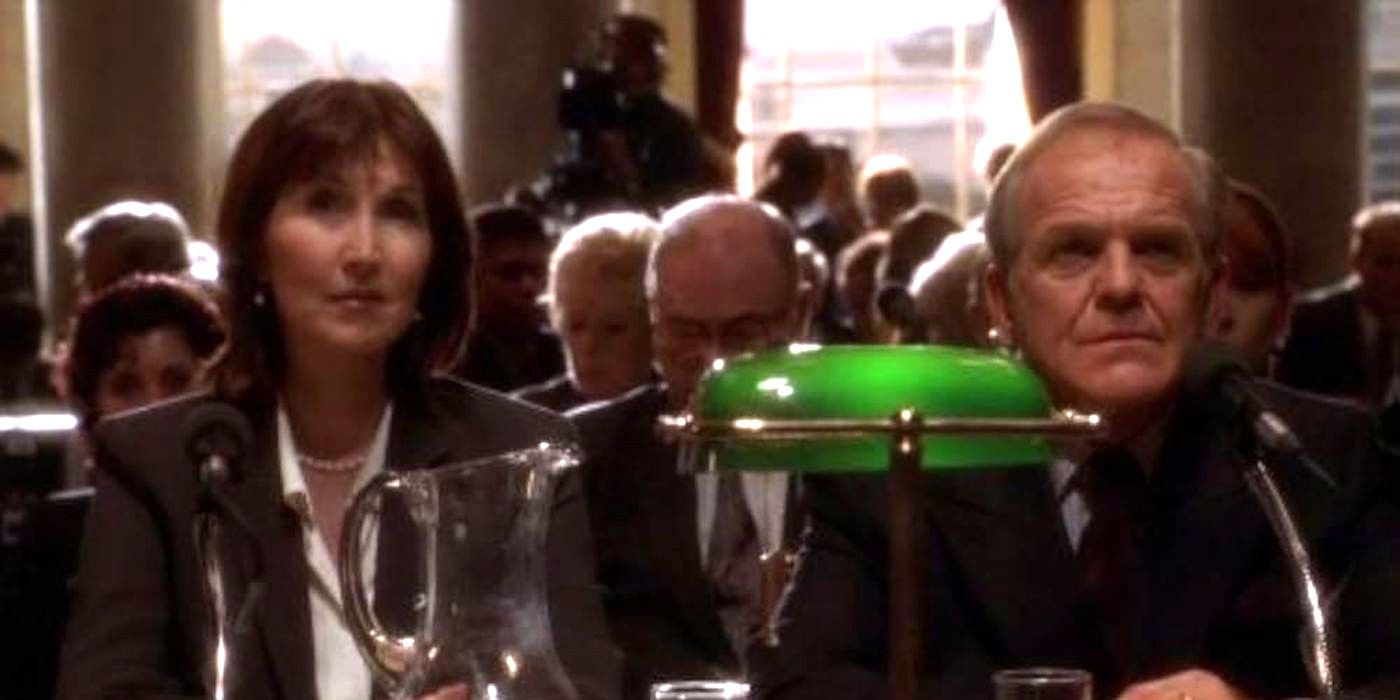 Leo Mcarry sits at a desk in front of a microphone in a room full of people and cameras in The West Wing.