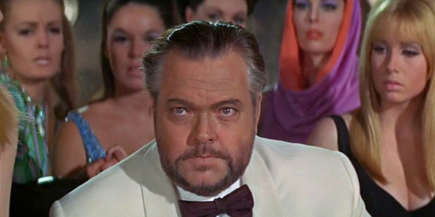 A man in a white tuxedo sits at a baccarat table surrounded by women in lavish and colorful dresses in 'Casino Royale' (1967).