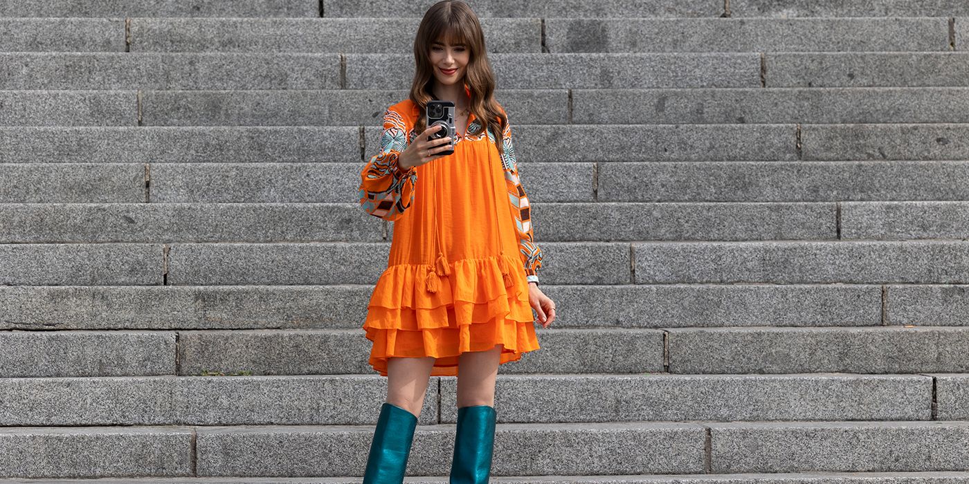 Emily from Emily in Paris standing on steps wearing an orange baby doll dress and taking a selfie.
