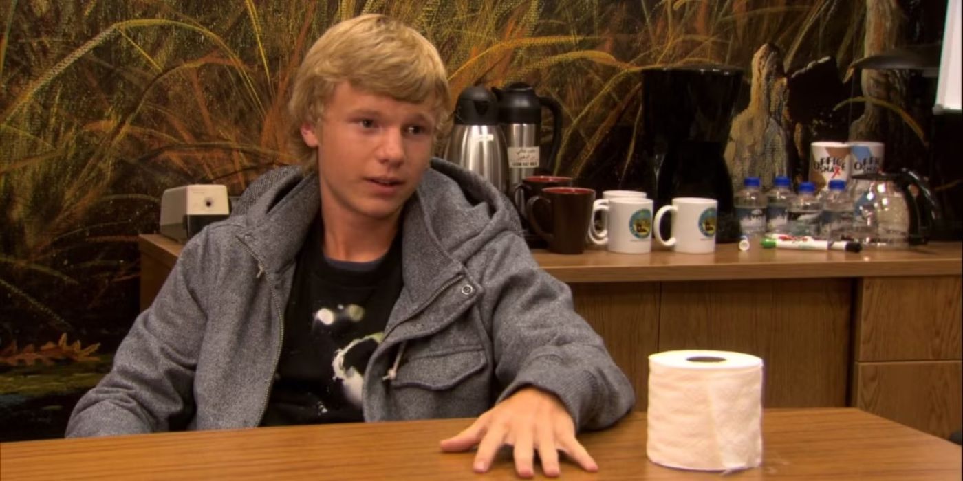 Teenager Greg Pikitis sits at a table with a roll of toilet paper and looks smug in Parks and Recreation.