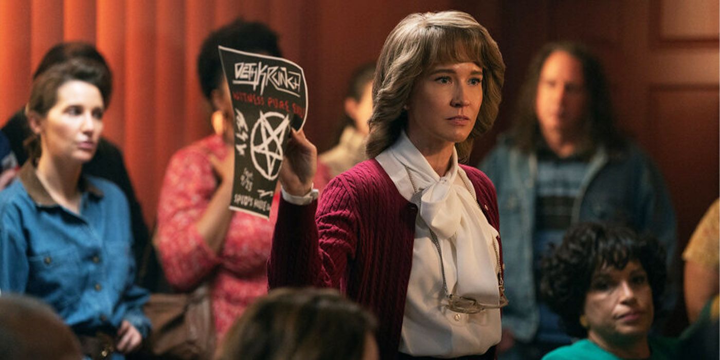 Anna Camp in '80s clothing and hairstyle holds up a satanic band poster in frustration