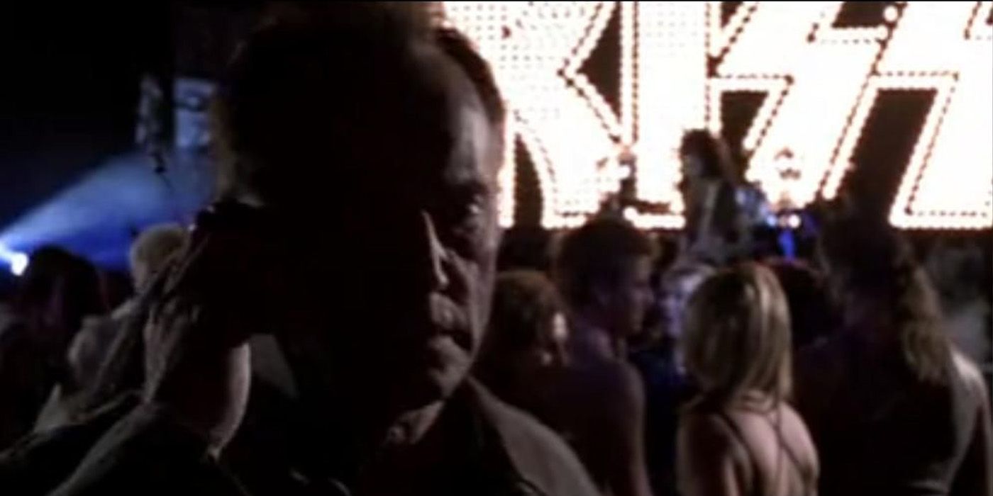 Lance Henriksen as Frank Black walks through a crowd as KISS performs on stage behind him