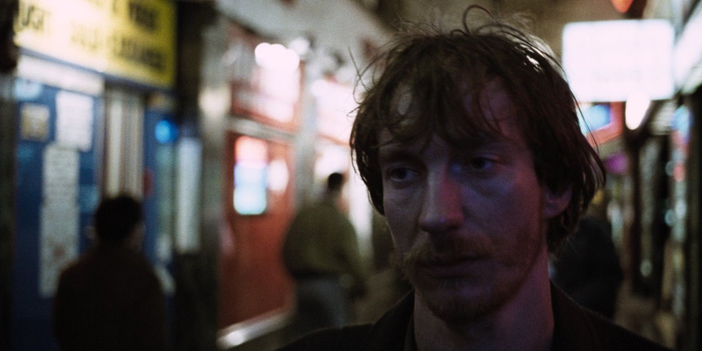 David Thewlis as Johnny looks blankly ahead walking down a corridor with people in the background in Naked.