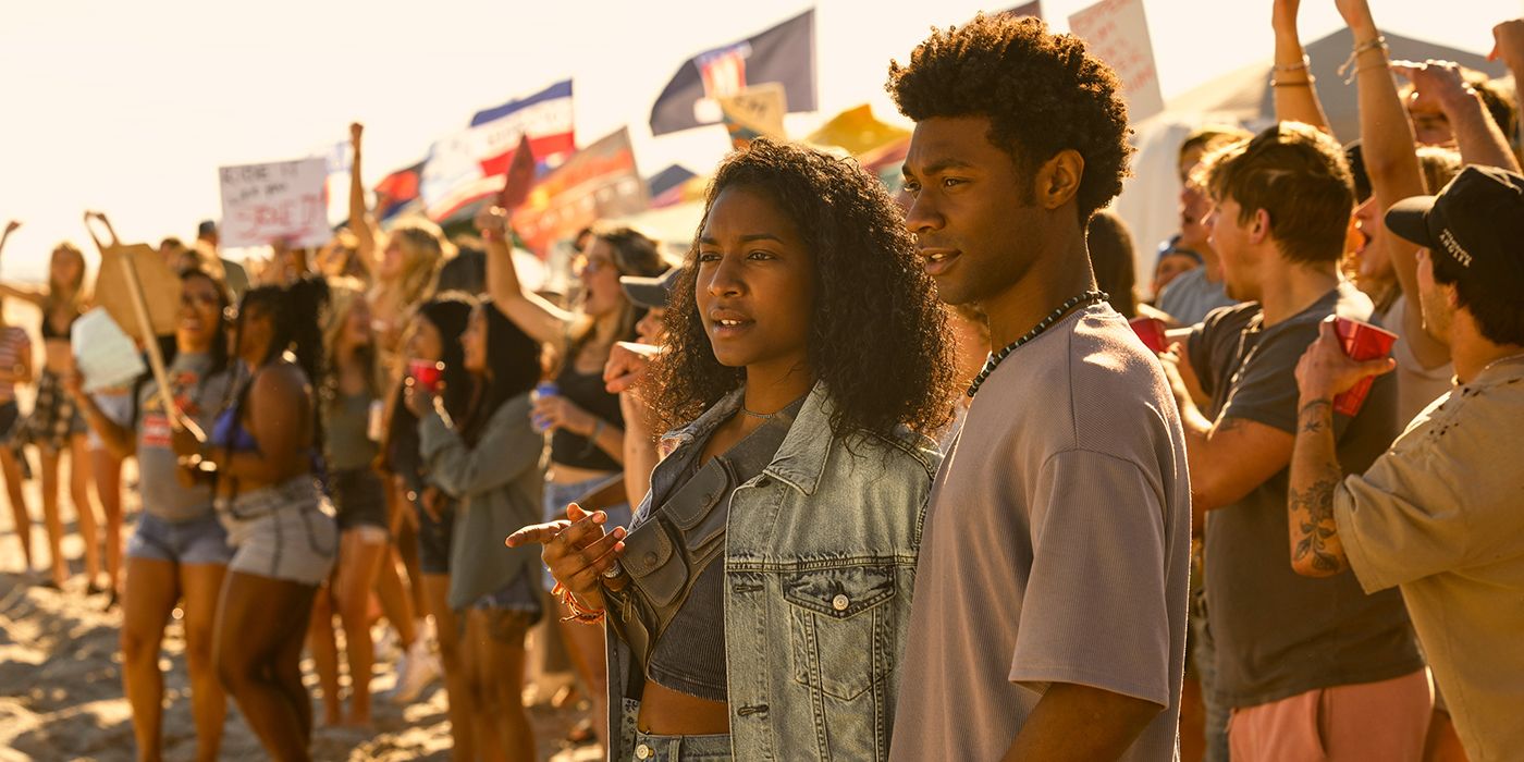 Carlacia Grant as Cleo and Jonathan Daviss as Pope standing with a crowd cheering on people during a race in Outer Banks Season 4 Part 1