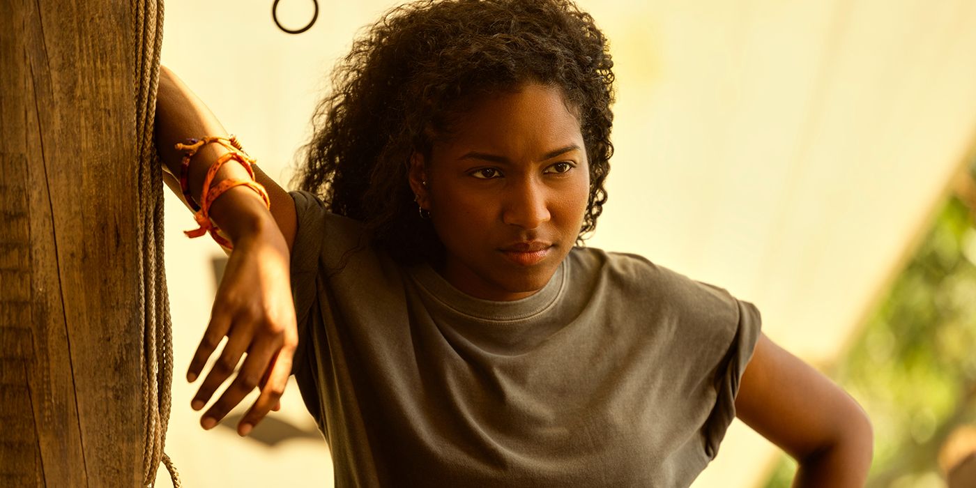 Carlacia Grant as Cleo resting her arm up against a wooden pole with an untrusting look on her face in Outer Banks Season 4 Part 1