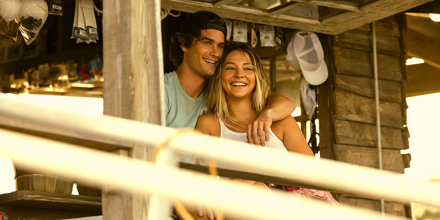 Chase Stokes with his arm over Madelyn Cline while standing on the dock looking out and smiling