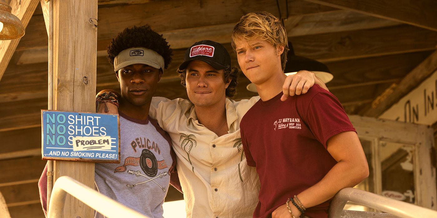 John B with his arms around Pope and JJ, standing together and looking out at someone in front of a sign that says no shirt, no shoes, no problem in Outer Banks Season 4 Part 1