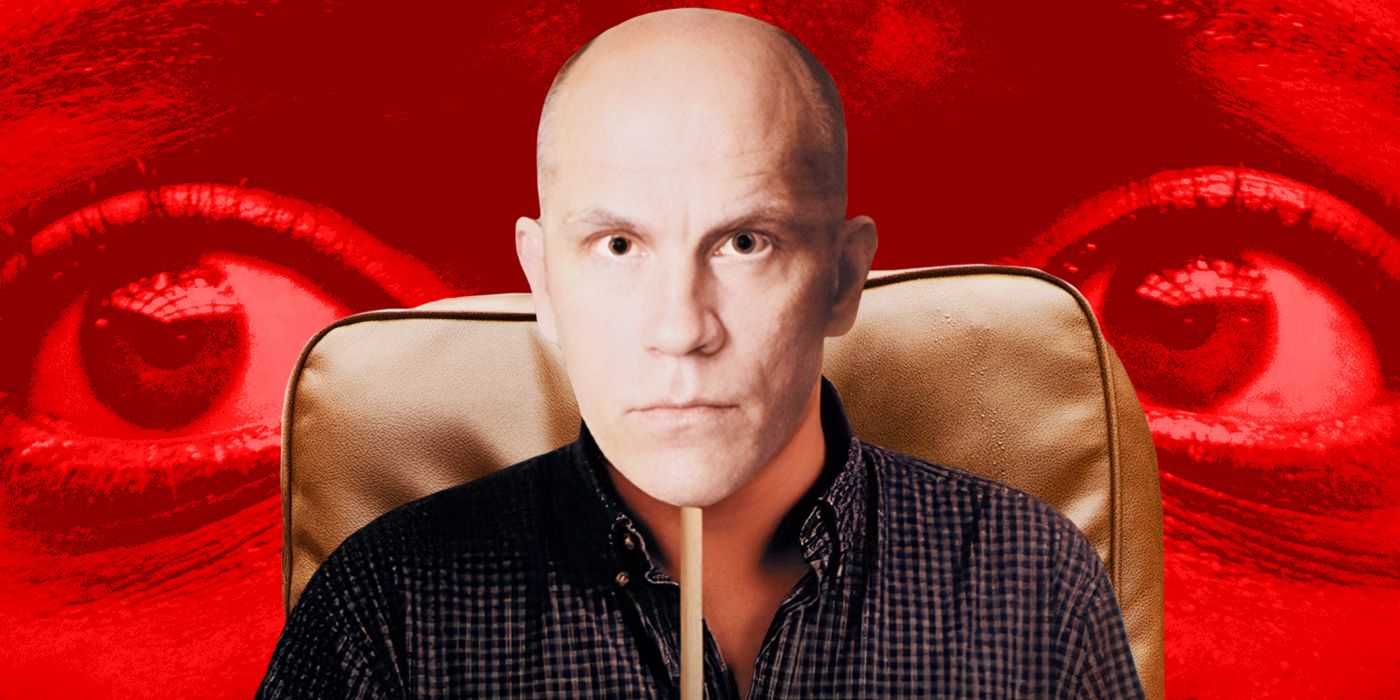 A person in a checked shirt sitting in a leather chair holding up a mask of actor John Malkovich.