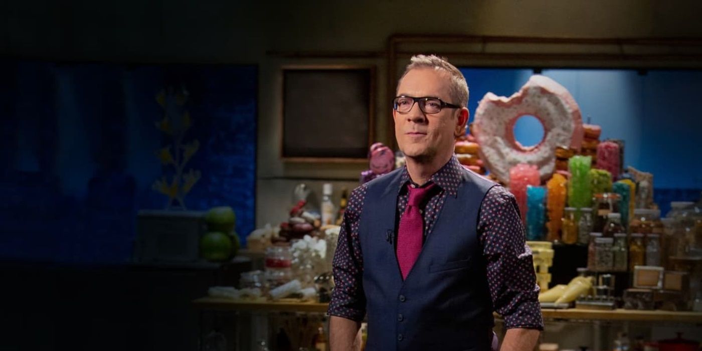 Ted Allen stands in front of a table full of sweets and a large doughnut on Chopped.