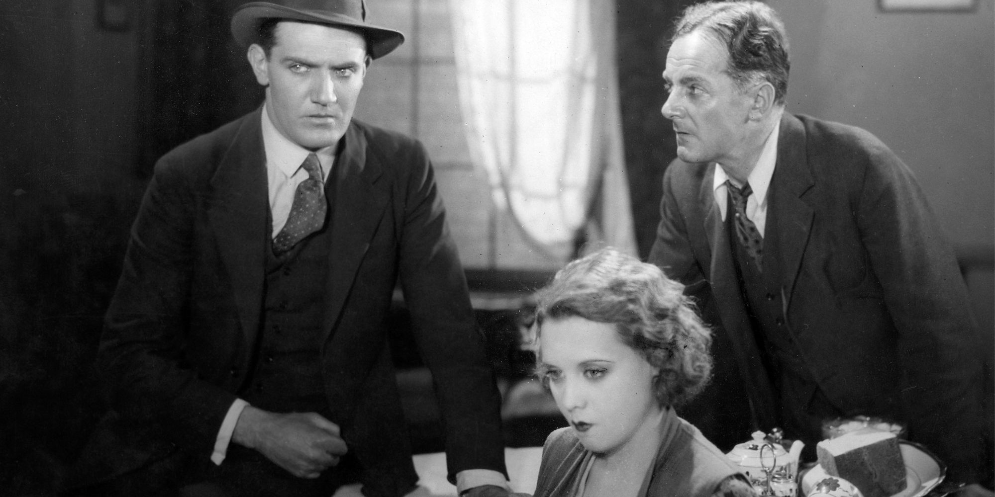 Donald Calthrop and John Longden standing on opposite sides of table while Anny Ondra sits in front of the table in Blackmail (1929)