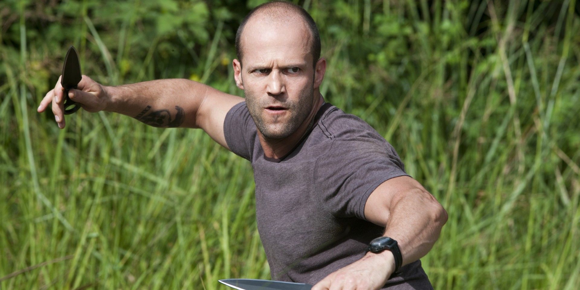 Jason Statham in tall grass wielding two knives in Expendables