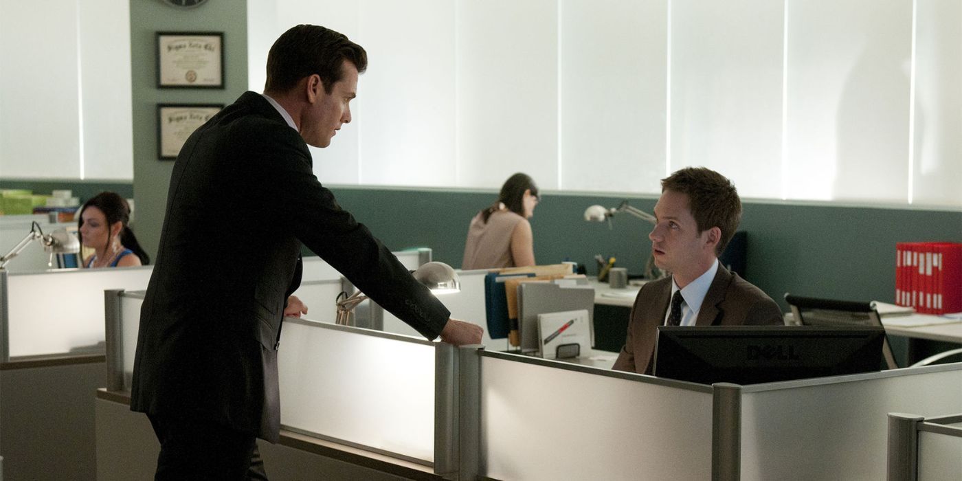 Gabriel Macht as Harvey Specter leans over a desk cubicle to talk to Patrick J. Adams as Mike Ross in Suits