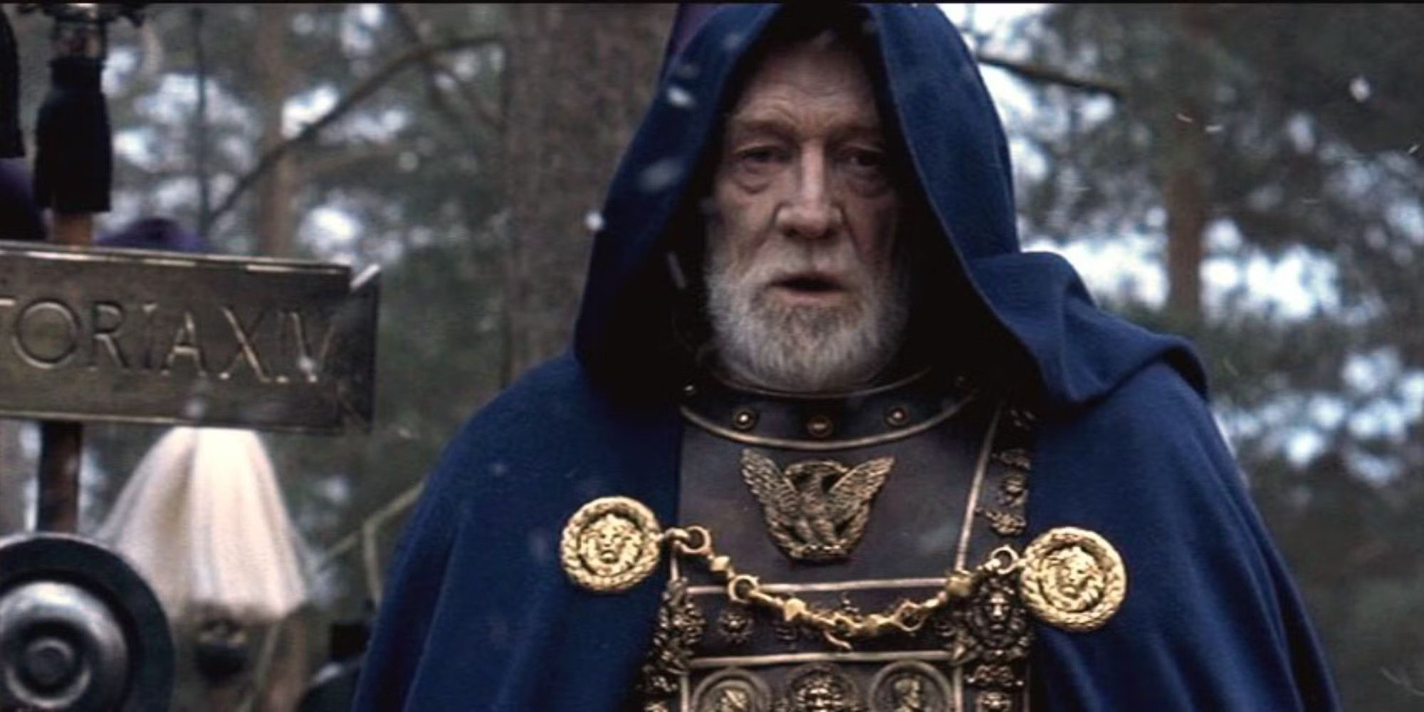 Richard Harris as Marcus Aurelius wearing a blue cloak and armor standing in a forest in Gladiator. 