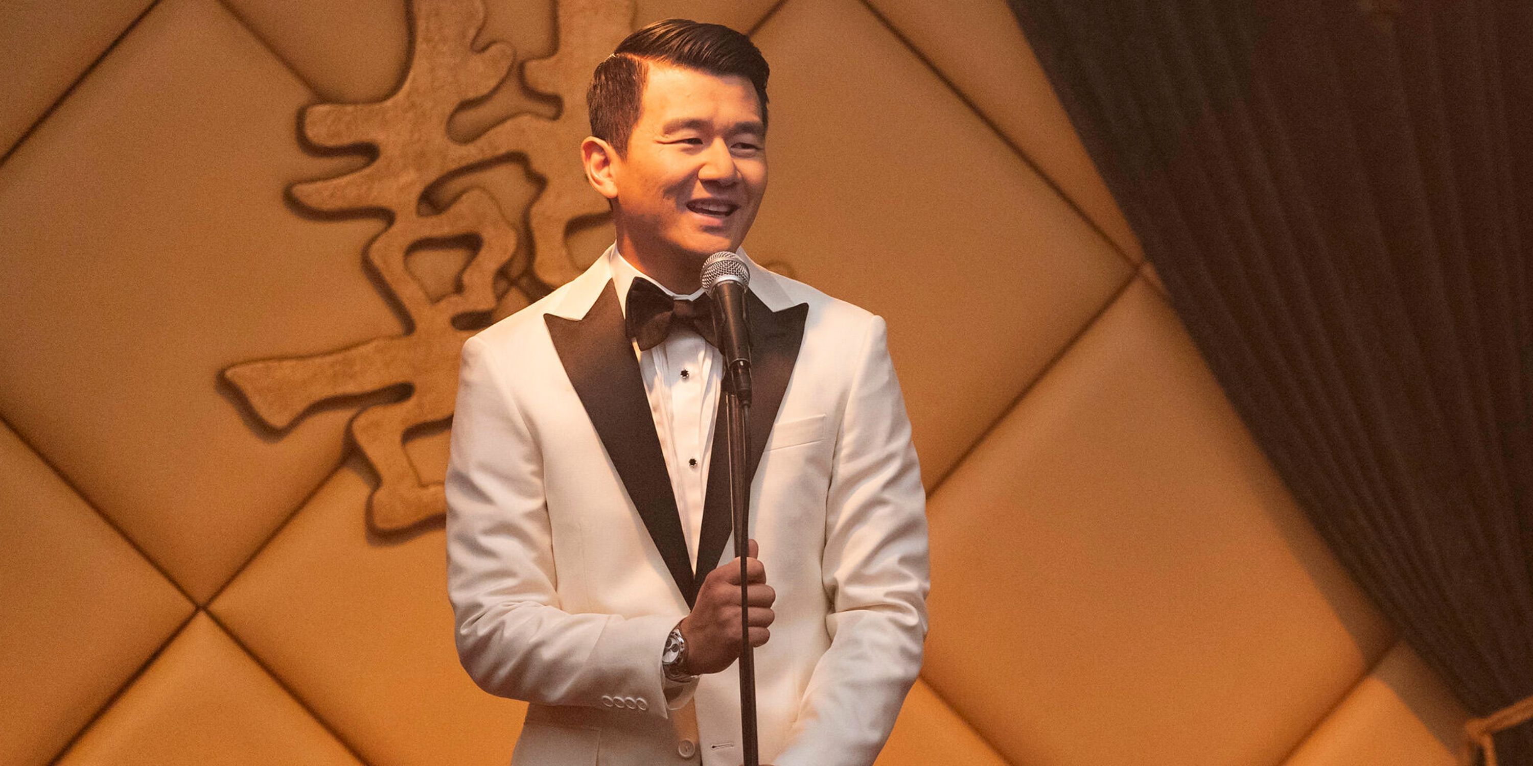Ronny Chieng as Fatty Choi in a white tuxedo standing on stage at a microphone in Interior Chinatown