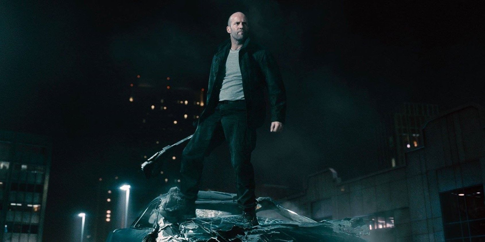 Jason Statham standing on top of a demolished car holding a weapon in Furious 7