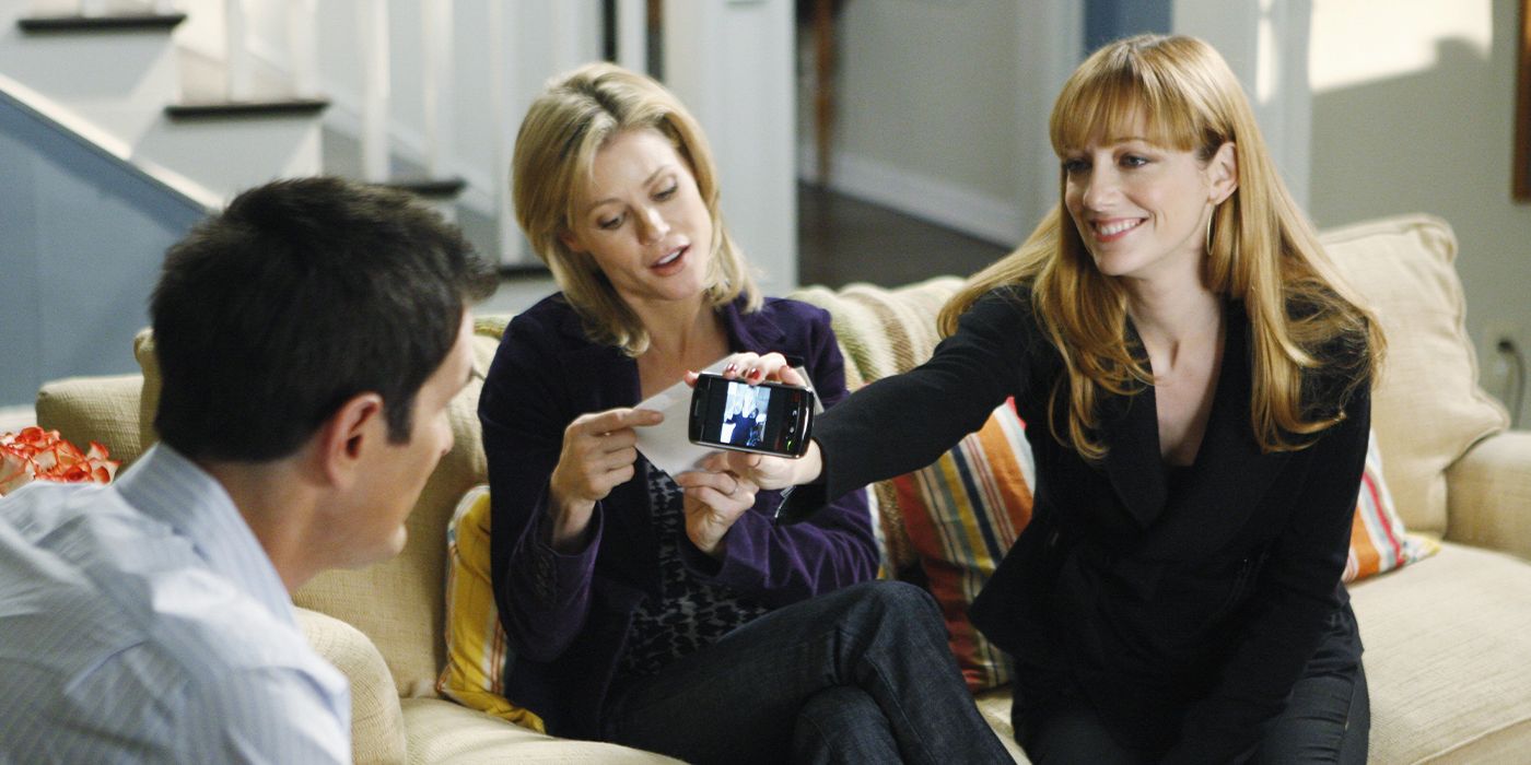 En Modern Family, Judy Greer se acerca a Julie Bowen, que está sentada en el sofá, y le muestra a Ty Burrell su teléfono celular.