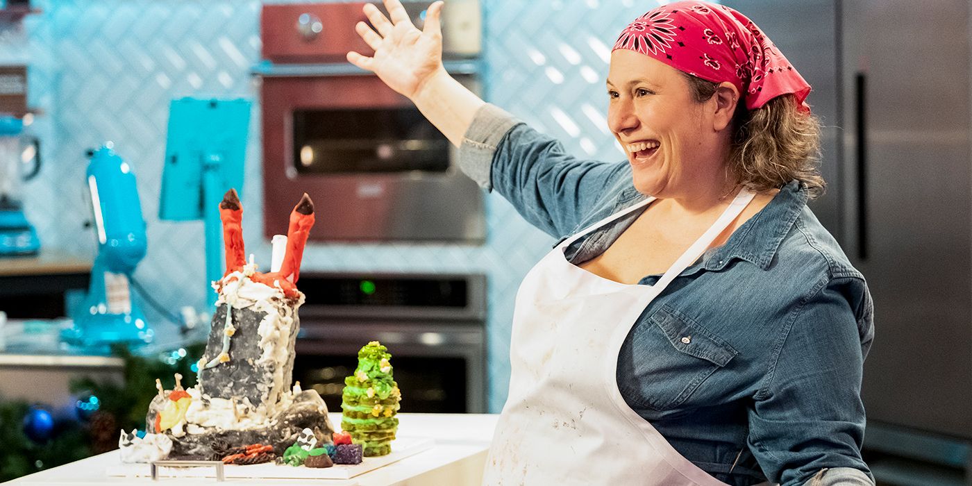 A woman spreads her arms in presentation and smiles at an awful cake creation in Nailed It! Holidays.