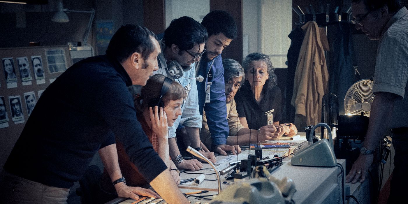 The cast leaning on a desk around Leonie Benesch with headphones on speaking into a microphone