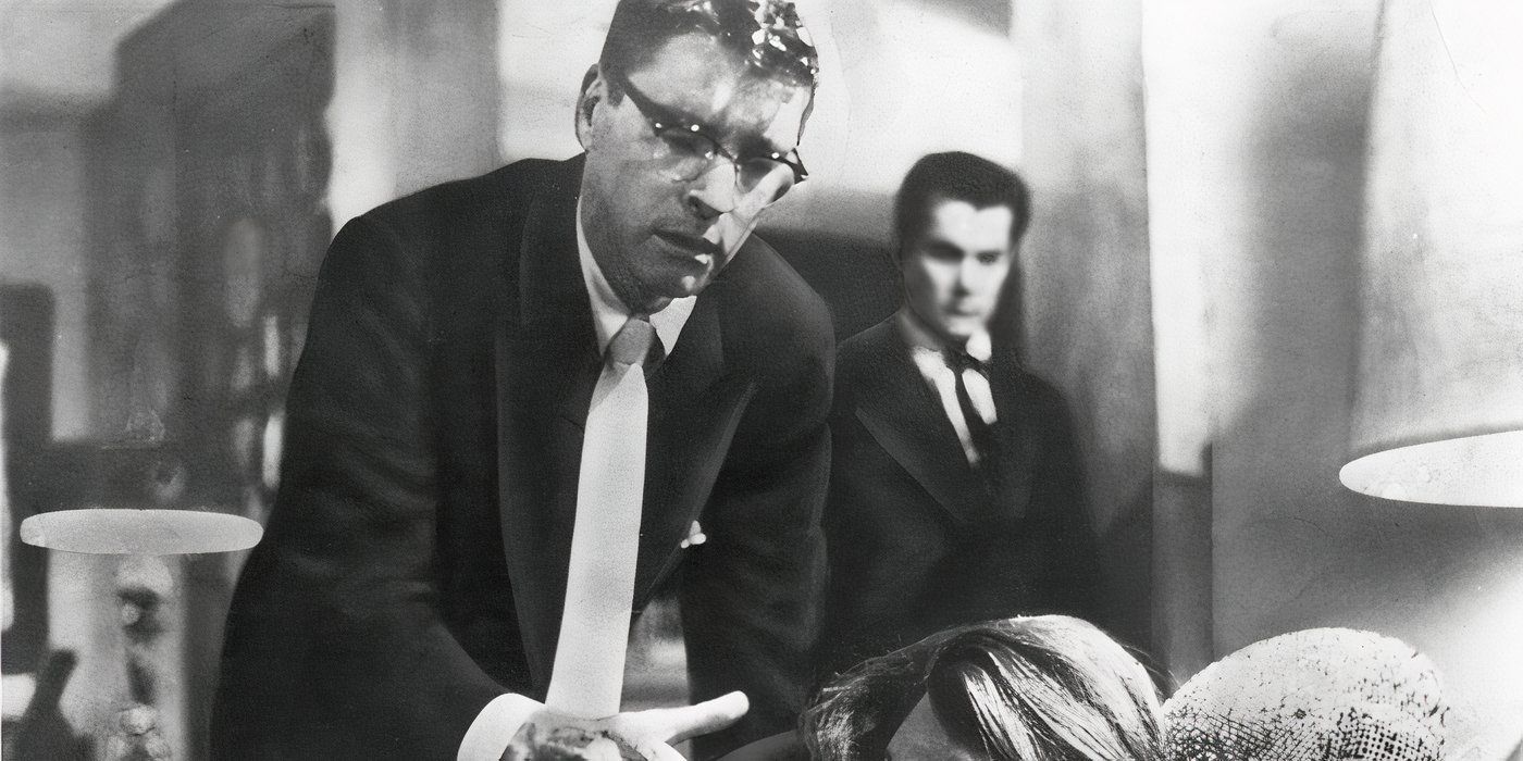 Two men in suits stand apart in a house with a person seated on a couch in front of them in Sweet Smell of Success